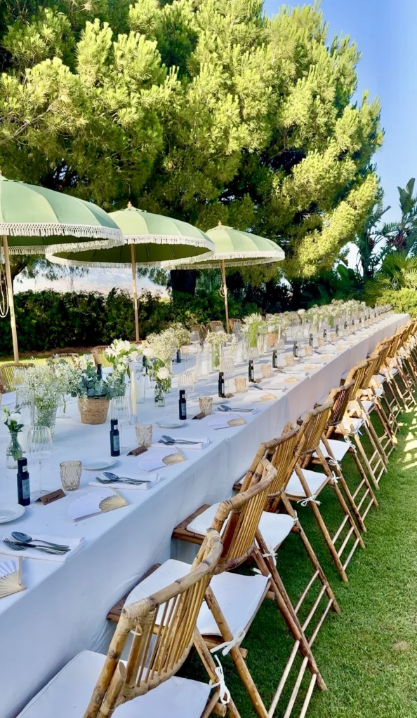 Long wedding banquet table with white linen, wooden folding chairs and green parasols in a garden near Marbella
