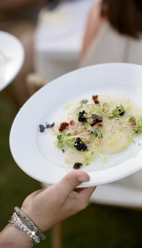 Hand holding a white plate of fish carpaccio garnished with edible flowers at a wedding in Marbella